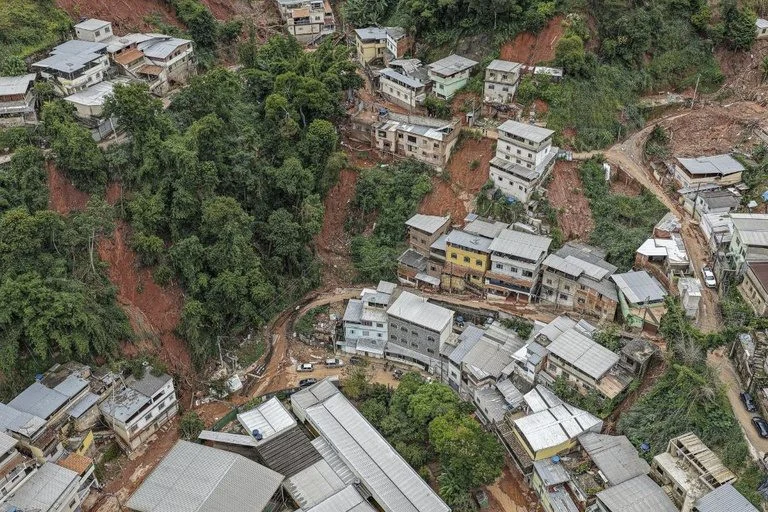 Imagem aérea mostra casas construídas em área de morro, cercadas por vegetação densa.Há trechos de encosta expostos, com solo avermelhado e sinais de deslizamento ou erosão.As moradias ficam próximas a barrancos e vias estreitas em uma região de ocupação irregular.