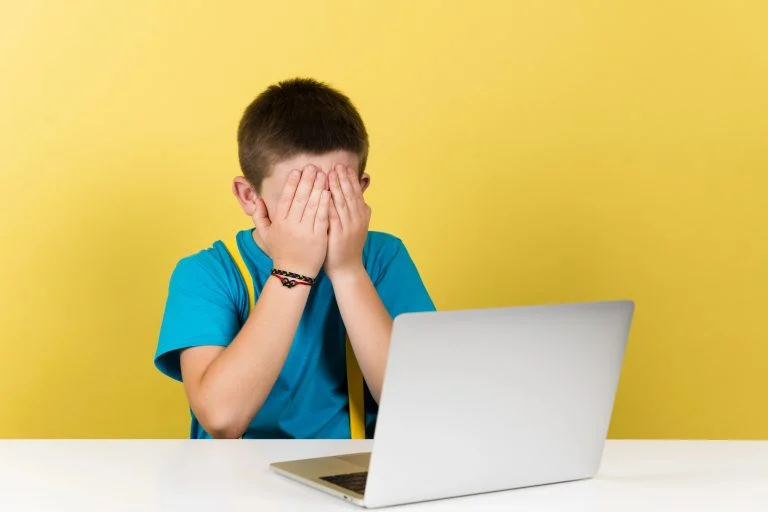 Menino com as mãos tampando os olhos e na frente um laptop