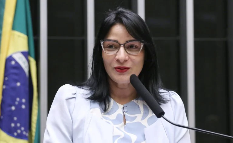Deputada Juliana Cardoso fala ao microfone. Ela é uma mulher clara, tem cabelos escuros e lisos na altura do ombro. Ela usa óculos e uma blusa clara. Atrás dela, à esquerda há uma bandeira do Brasil