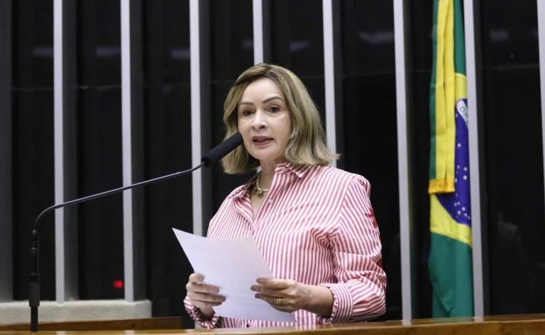 Deputada Socorro Neri fala ao microfone. Ela é uma mulher branca, tem o cabelo liso, loiro, na altura dos ombros, veste uma camisa listrada de vermelho e branco e segura um papel
