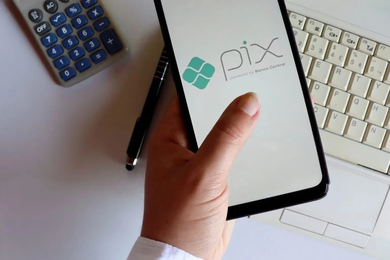 Uma mão segura um celular que exibe o logotipo do Pix, sistema de pagamentos do Banco Central. Ao redor, sobre uma mesa clara, há uma calculadora, uma caneta e parte do teclado de um computador.
