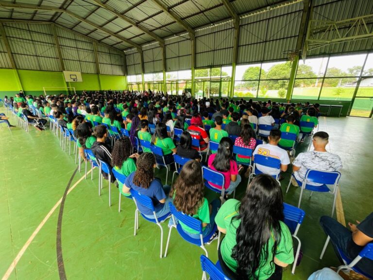 Alunos da escola sentados em cadeiras na quadra esportiva coberta, de frente para o palco com o professor empunhado de um violão.