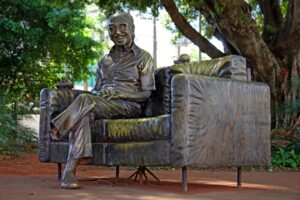 Estatua de Manoel de Barros. Escritor está sorrindo, sentando, com as pernas cruzadas. O pé esquerdo dele está faltando.