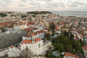 Imagem aérea mostra o casario colorido de Lisboa com destaque para um edifício branco com uma torre e uma piscina redonda, onde fica o hotel Torel Palace Lisboa